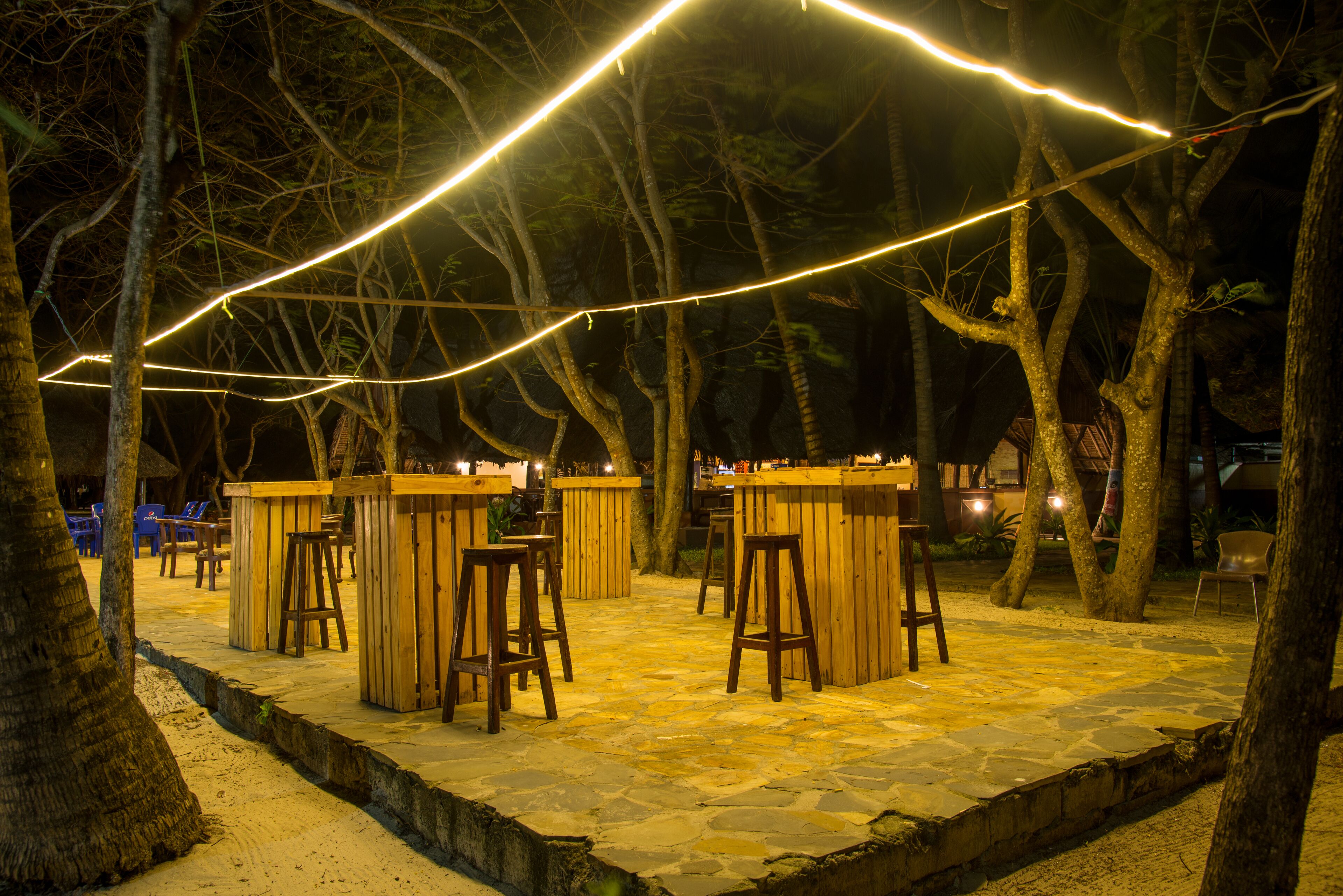 An outdoor bar area with wooden stools and tables, surrounded by trees and illuminated by string lights.