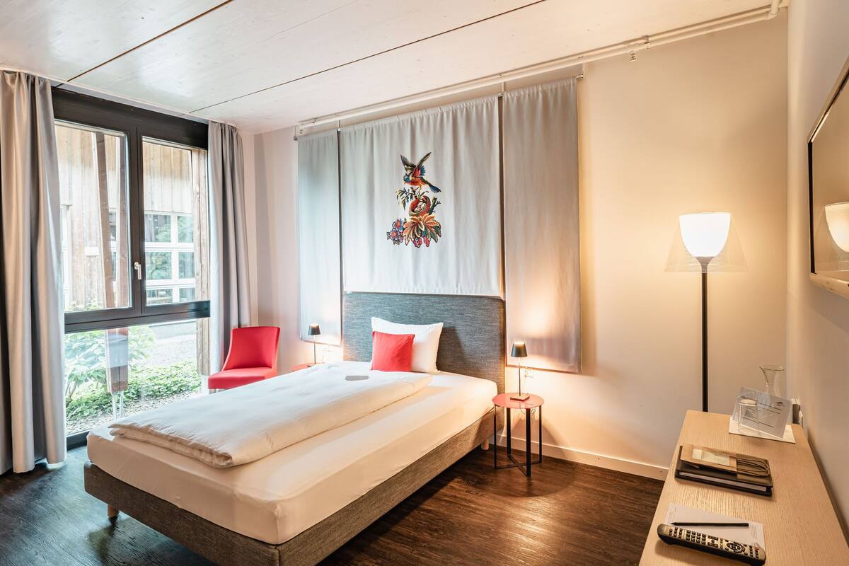 A hotel room with a bed, a red chair, a desk with a lamp, and a window with curtains.