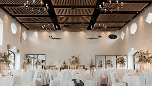 Une vaste salle dotée de hauts plafonds, de poutres en bois et de lustres. Des tables sont dressées avec des nappes blanches et des chaises, et des compositions florales ornent les tables.