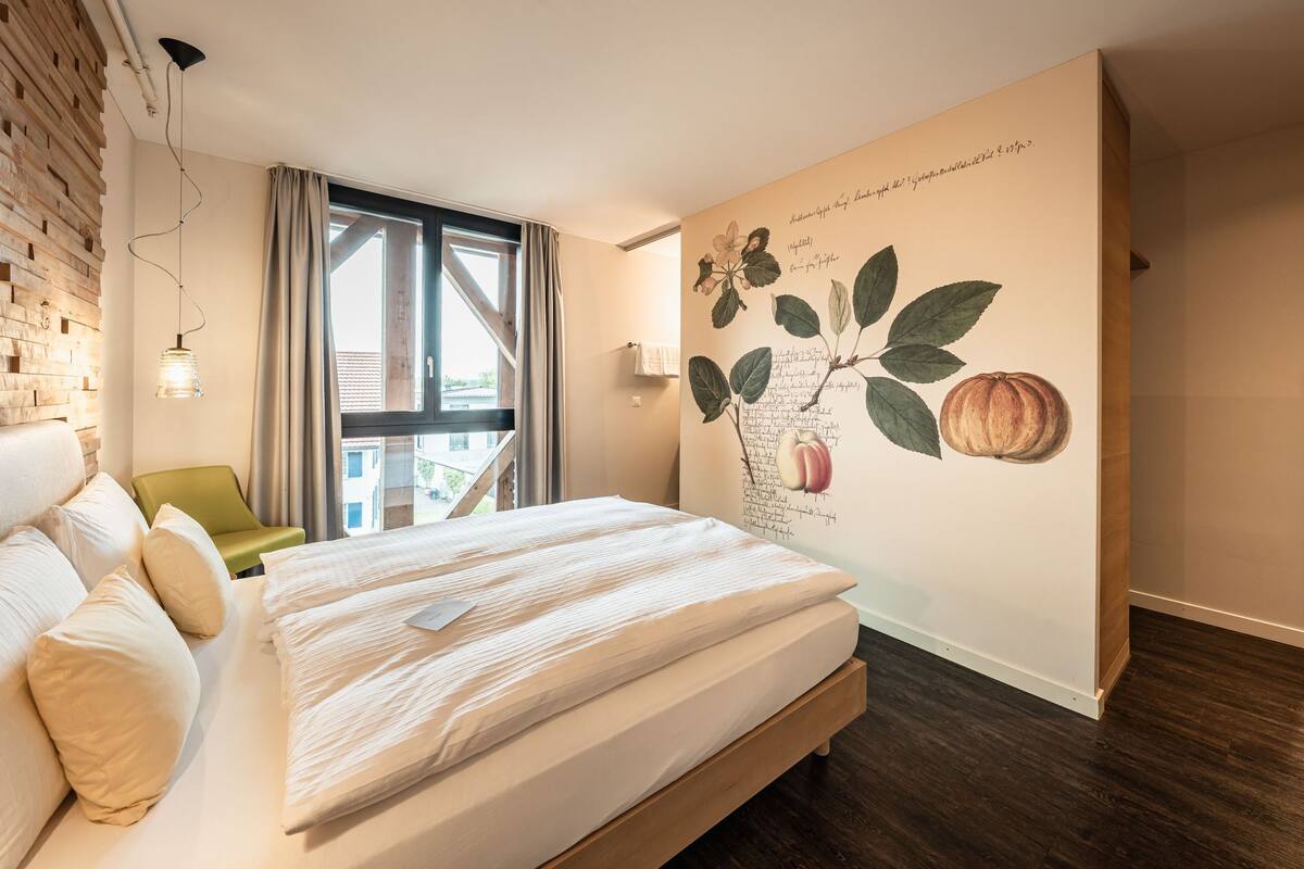 A modern bedroom with a large bed, a green chair, a window with curtains, and a wall mural of fruit and text.