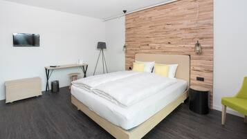 A modern bedroom with a wooden headboard, a bed with white bedding, a desk with a chair, a standing lamp, and a television on the wall.