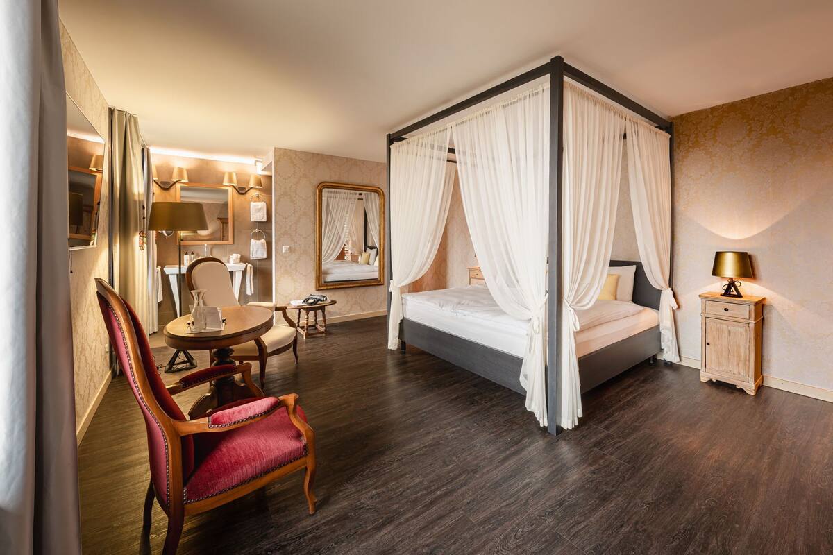 A four-poster canopy bed with white drapes, a wooden chair with red upholstery, a bedside table with a lamp, and a mirror on the wall.
