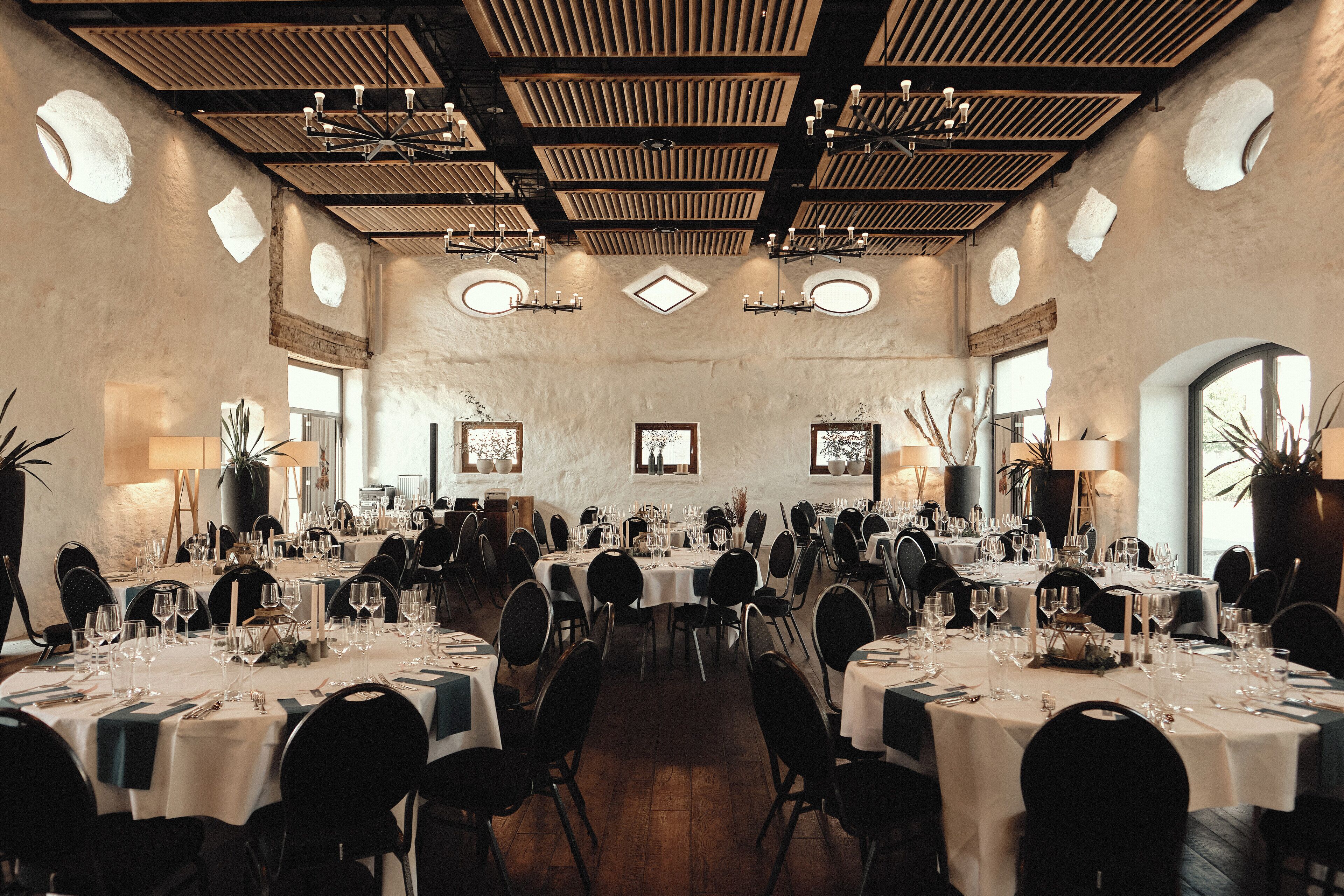 A large dining hall with multiple tables set for a formal event, featuring a high ceiling with wooden beams and circular skylights.
