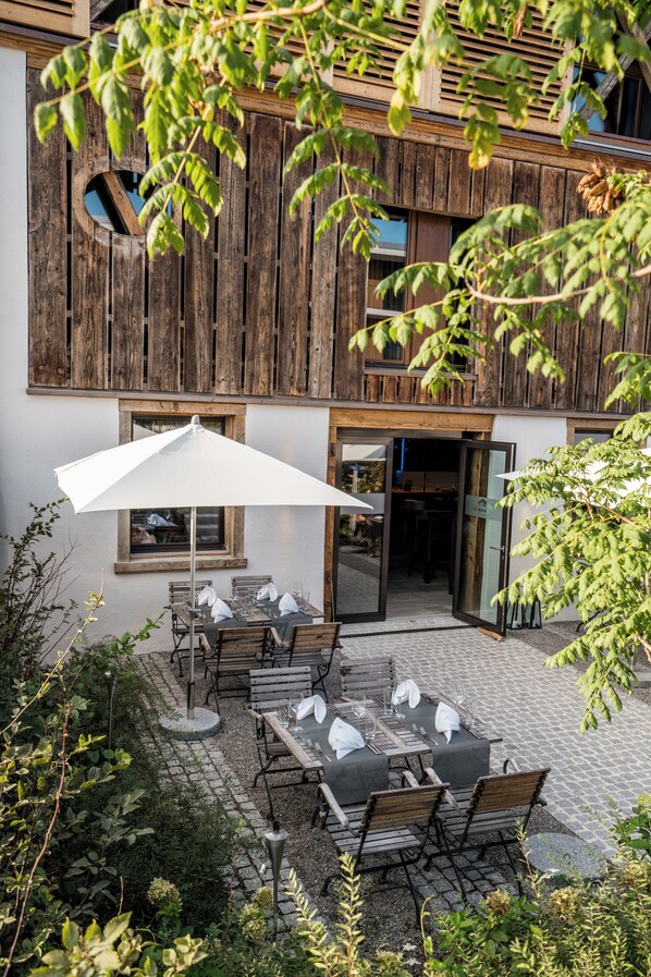 Un patio al aire libre con mesas y sillas, una sombrilla blanca y un edificio de madera con aspecto rústico.