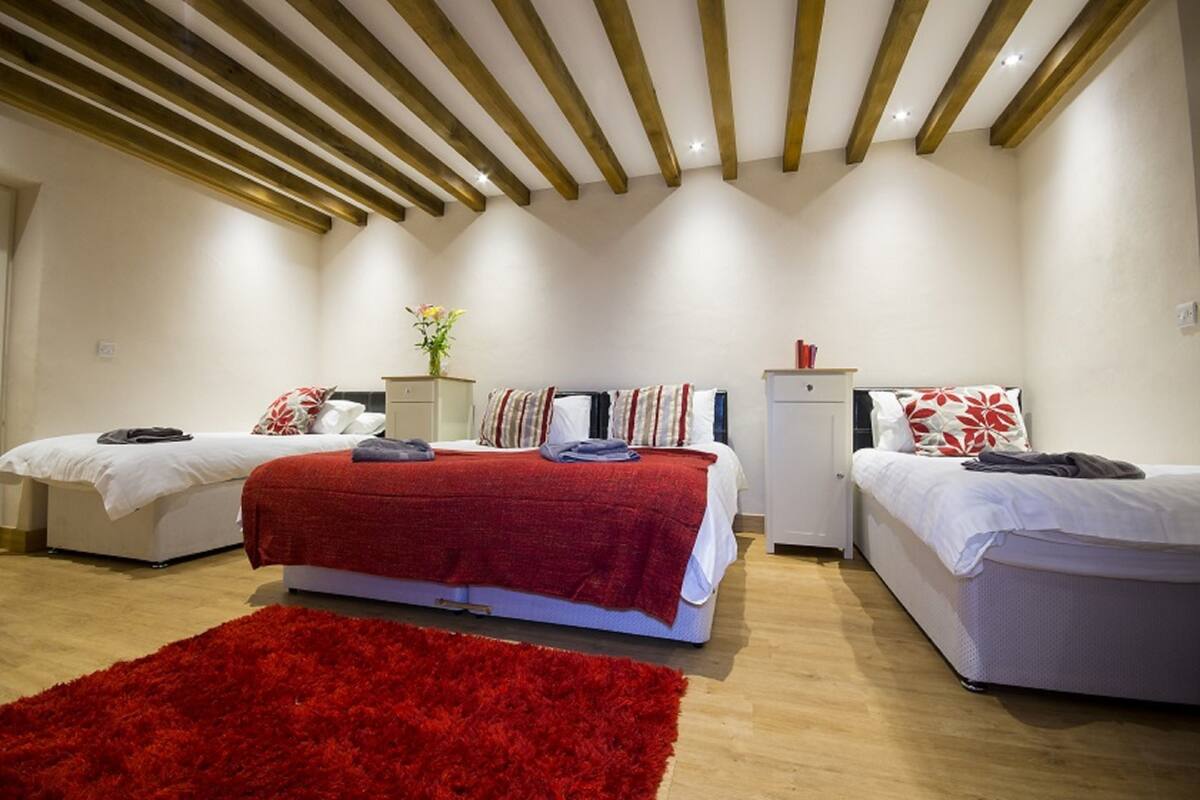 Une chambre avec deux lits, un tapis rouge, des poutres en bois au plafond et une petite armoire blanche.