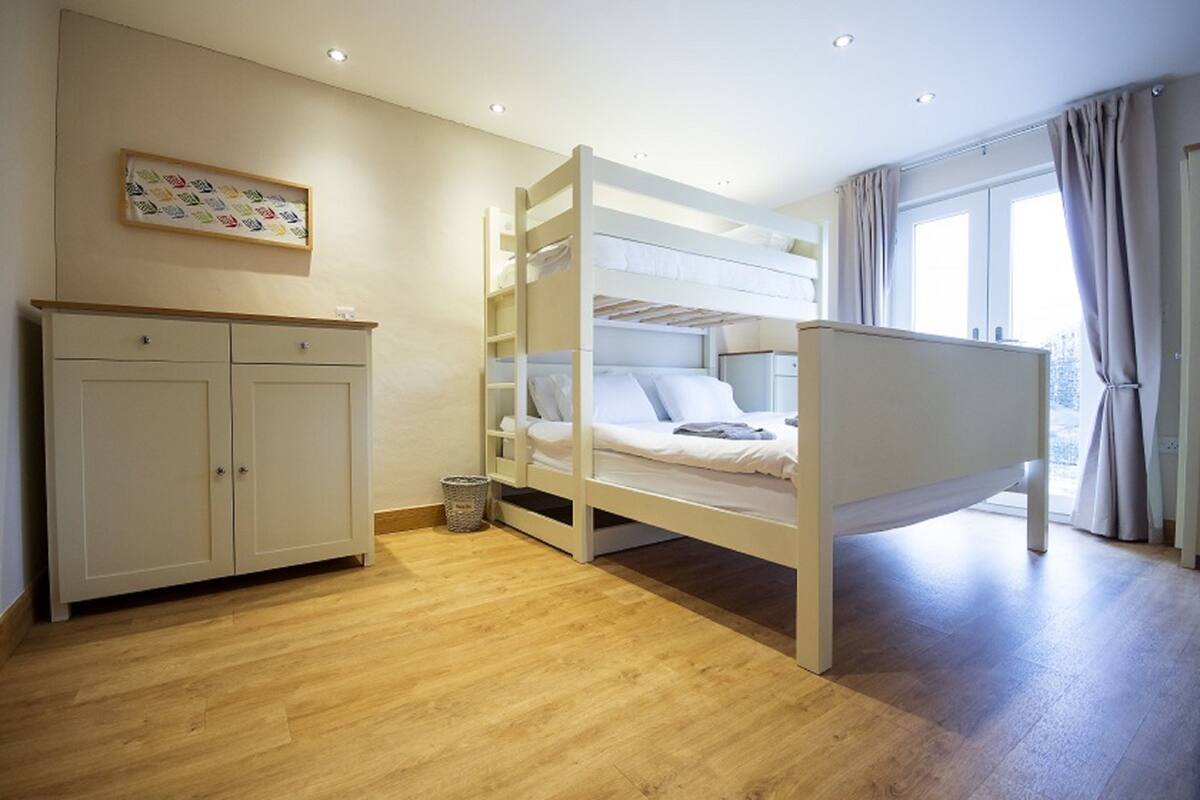 Un lit superposé avec une structure en bois, une armoire blanche et une petite poubelle dans une pièce dont le sol est recouvert de planches de bois et qui possède une fenêtre avec des rideaux.