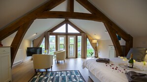 A bedroom with a sloped ceiling, a bed with a tray of champagne, a bedside table, a chair, and a view of the outdoors. - Tynrhyd Retreat (Aberystwyth)