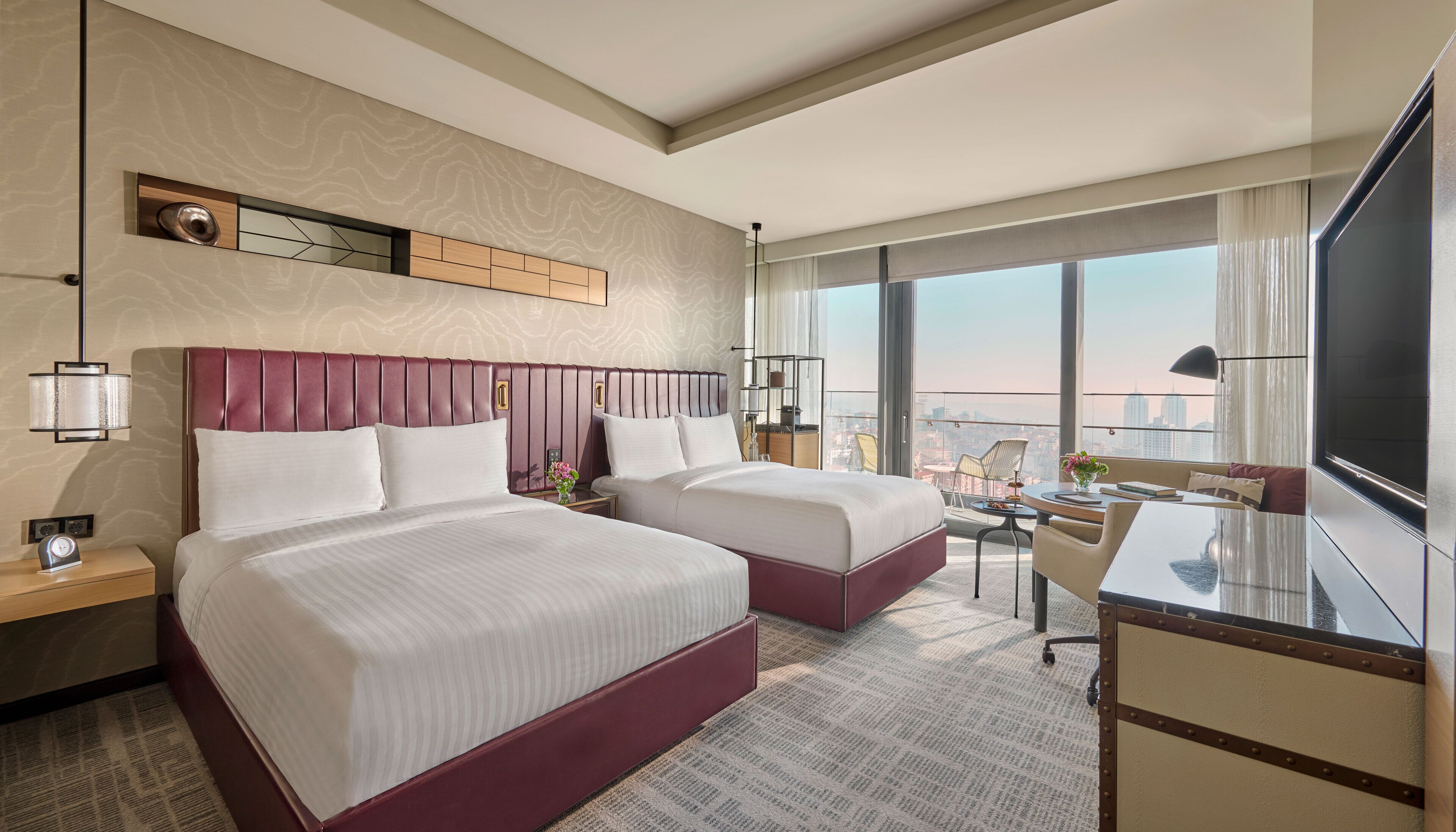 A hotel room with two beds, a desk, a chair, a TV, and a large window with a city view.