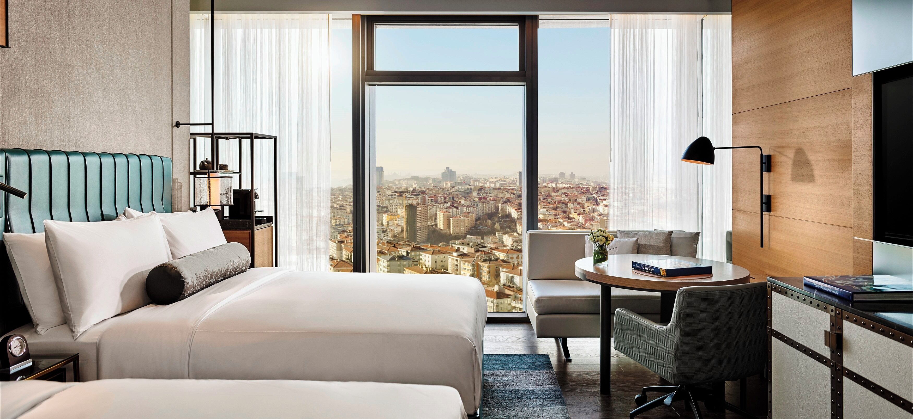 A hotel room with a large window offering a city view, a bed with a headboard, a desk with a chair, and a TV on the wall.