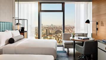 A hotel room with a large window offering a city view, a bed with a headboard, a desk with a chair, and a TV on the wall.