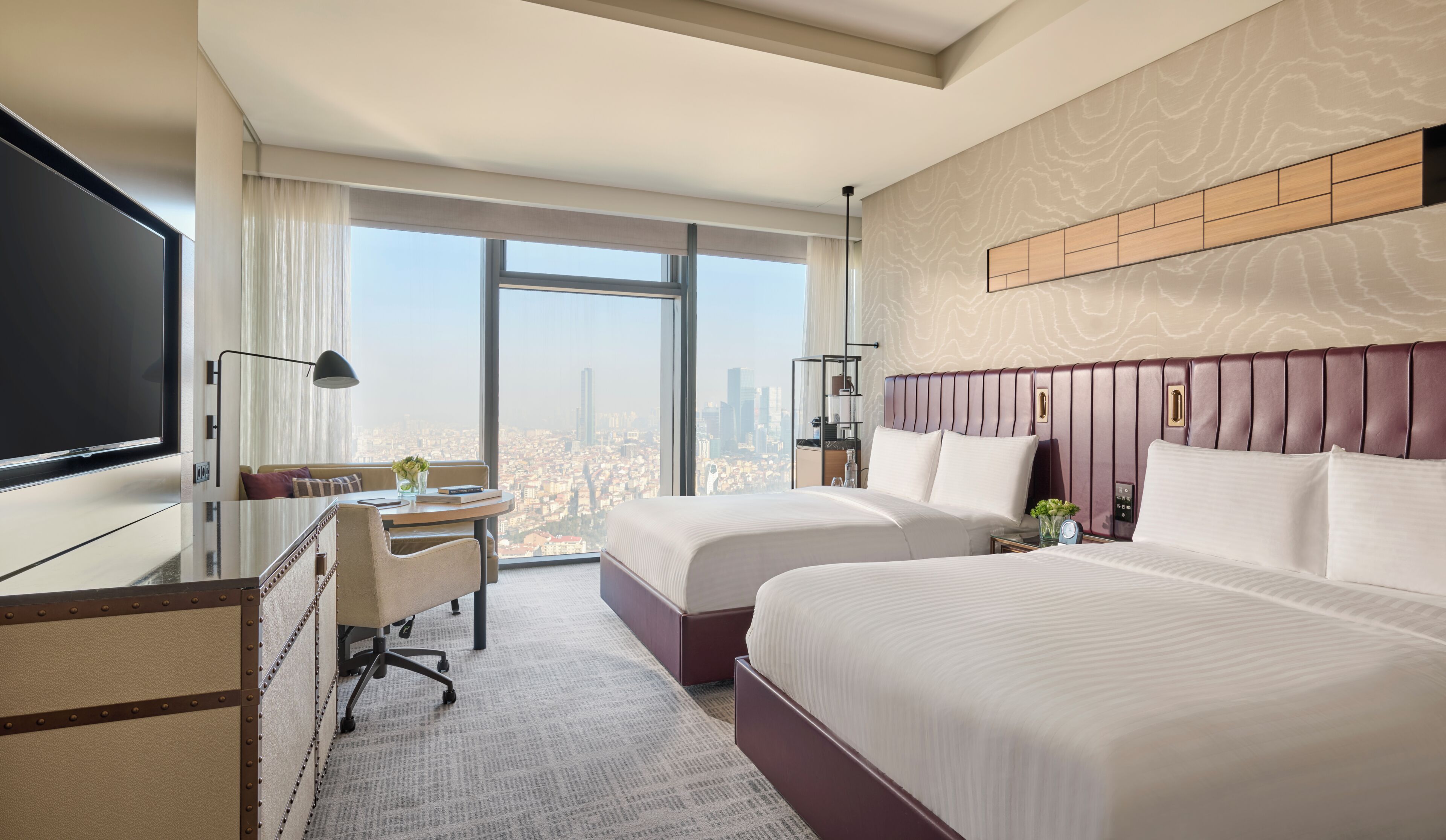 Ein Hotelzimmer mit zwei Betten, einem Schreibtisch, einem Fernseher und einem großen Fenster mit Blick auf die Stadt.