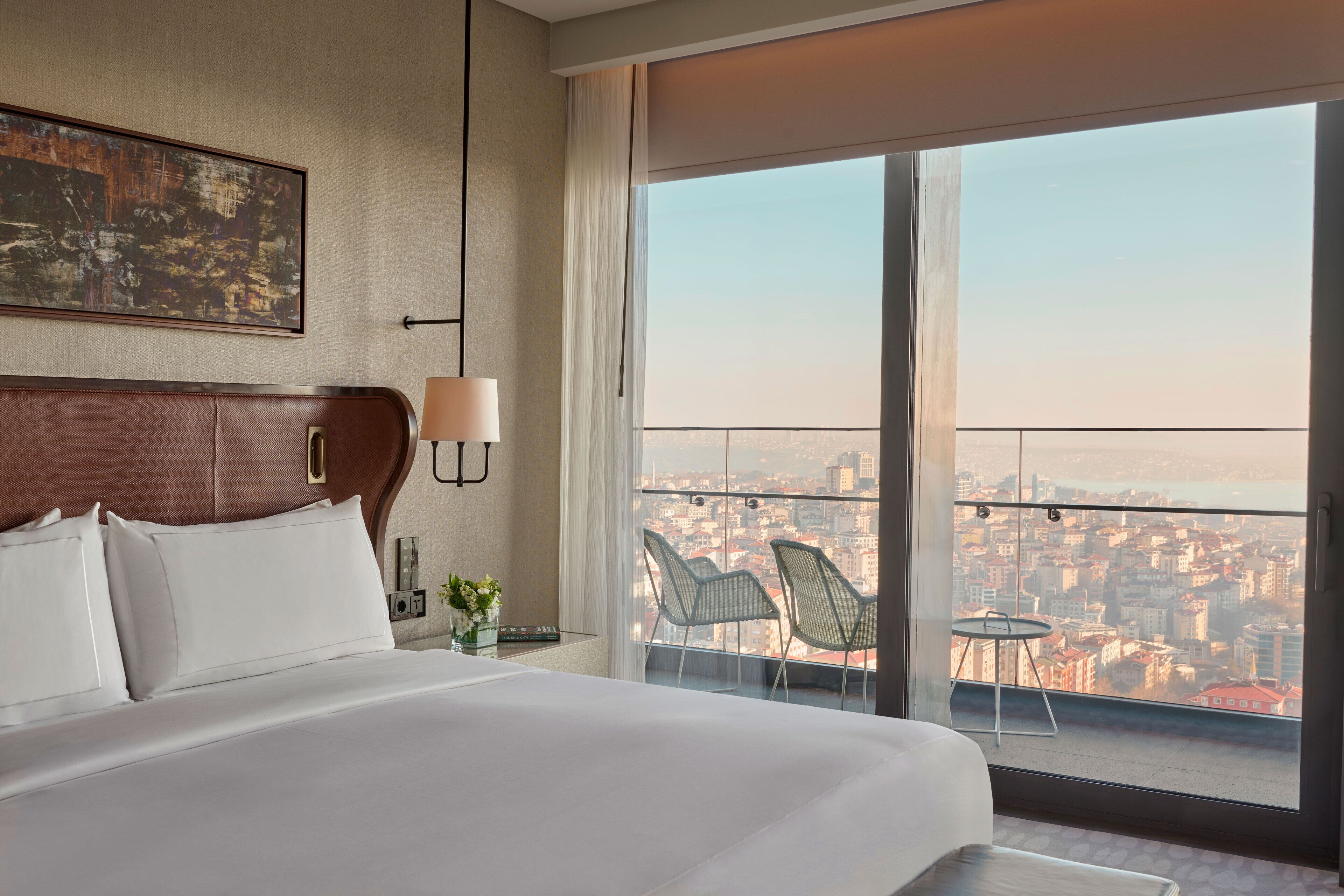 A hotel room with a large bed, a city view through the window, and a small table with chairs.