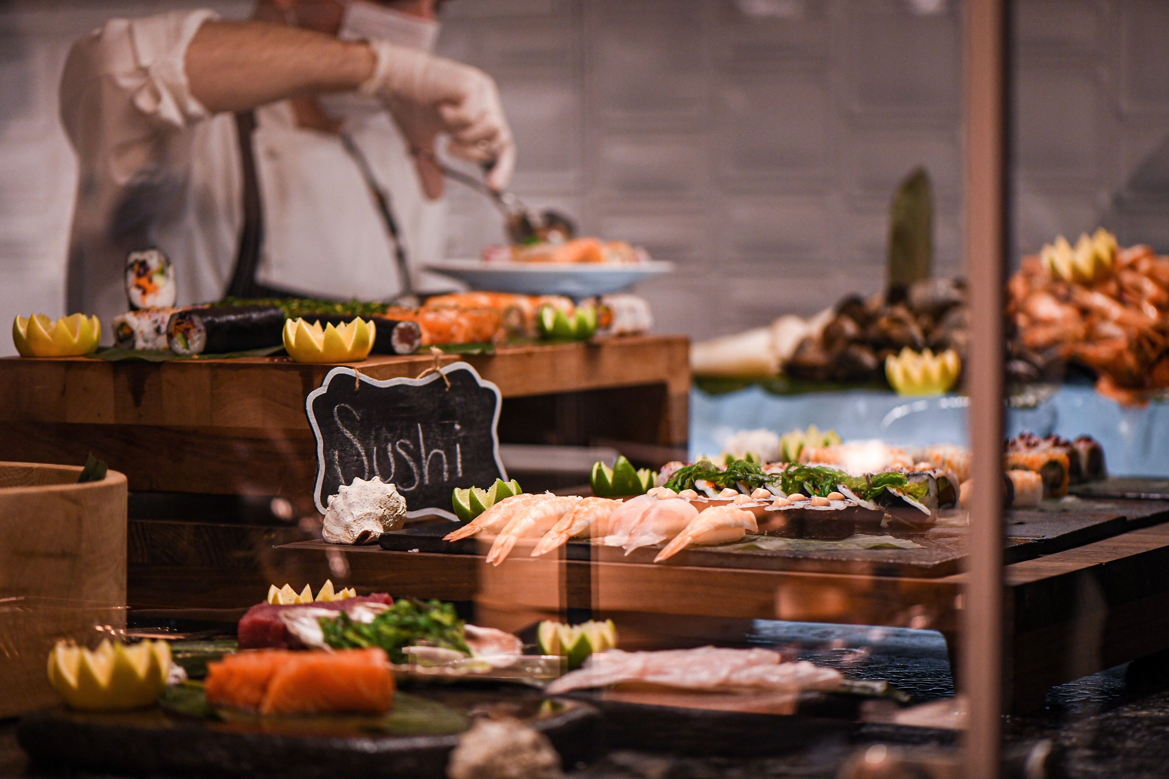 A sushi bar with various types of sushi displayed on wooden tables, a chalkboard sign reading "Sushi", and a chef preparing food in the background.