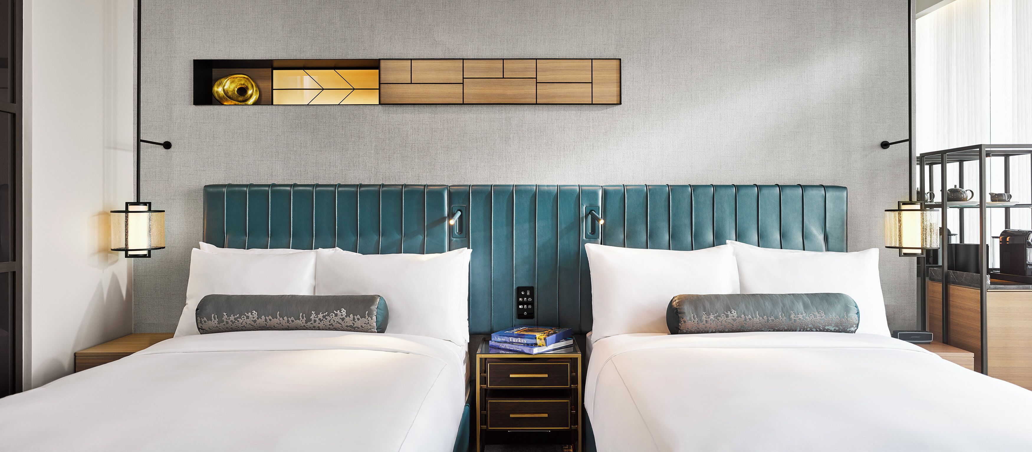 A hotel room with two beds, a wall-mounted light fixture, a small table with a lamp, and a shelf with a teapot.