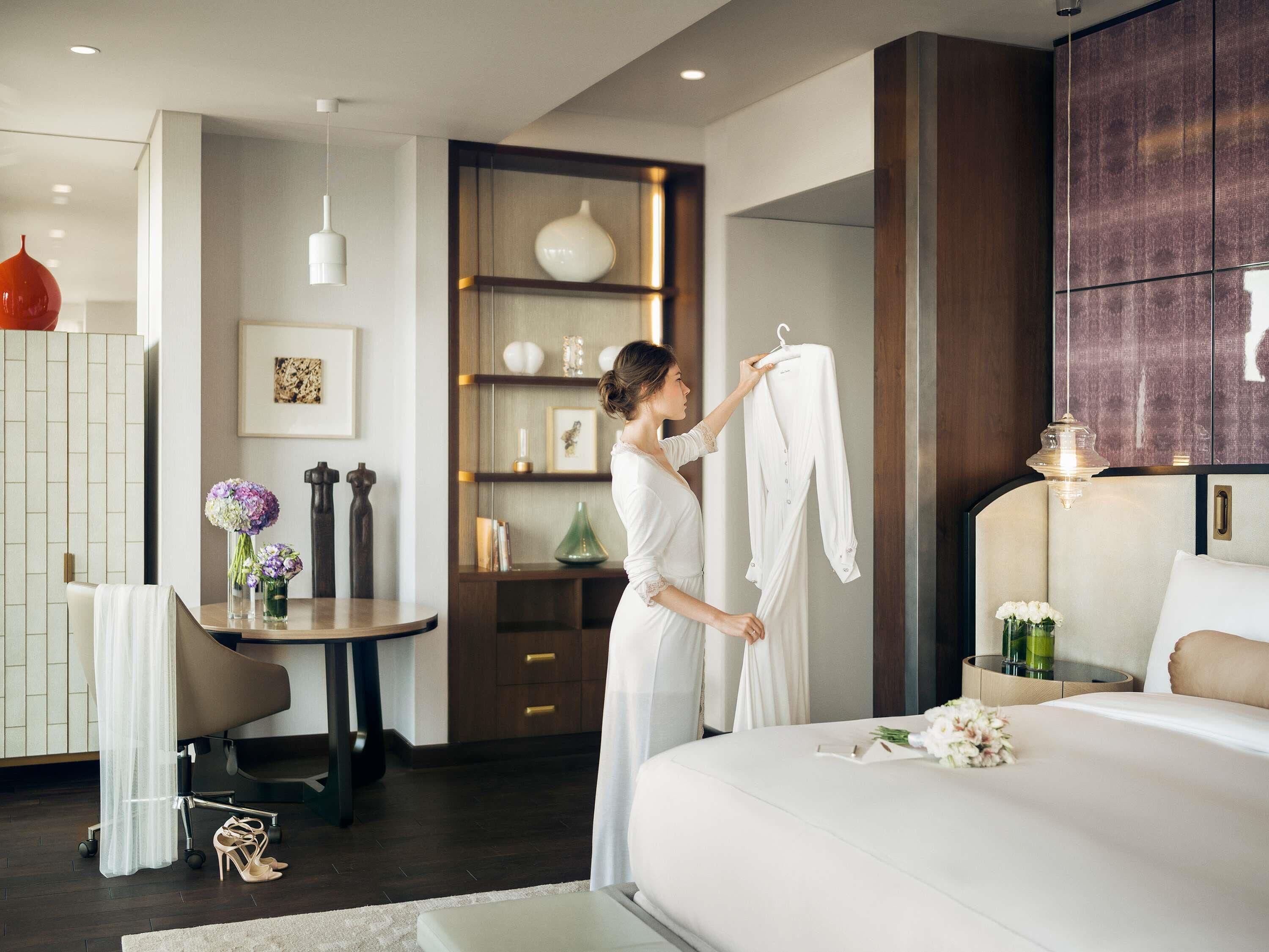 A modern hotel room with a large bed, a bedside table with a lamp, a small desk with a chair, and a decorative shelf with various items.