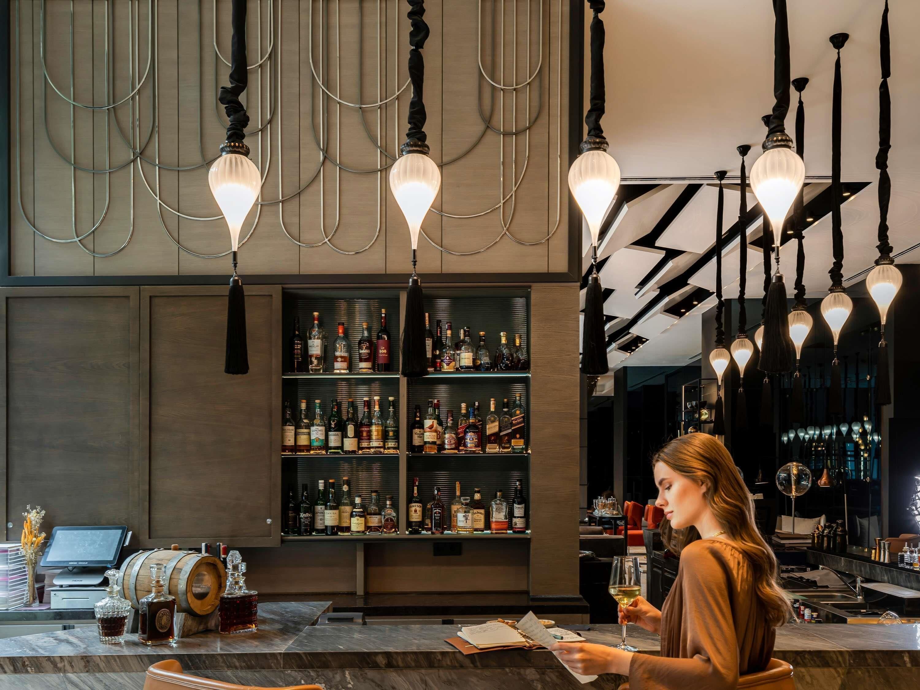 Zona de bar con estanterías llenas de botellas de licor, un barril de madera y una persona sosteniendo una copa.