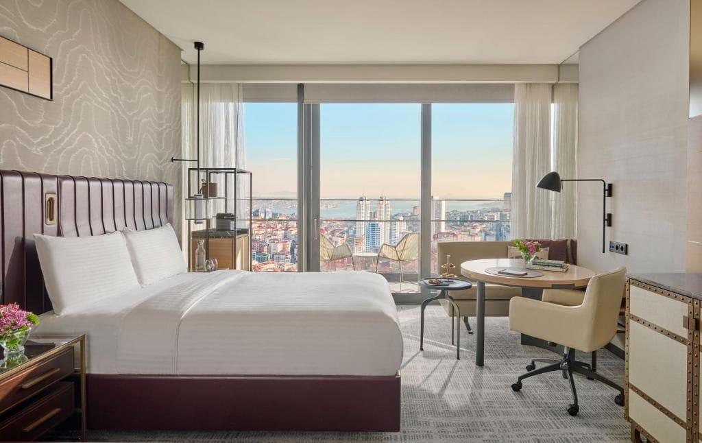 A hotel room with a large bed, a desk, a chair, a small table, and a view of the city.