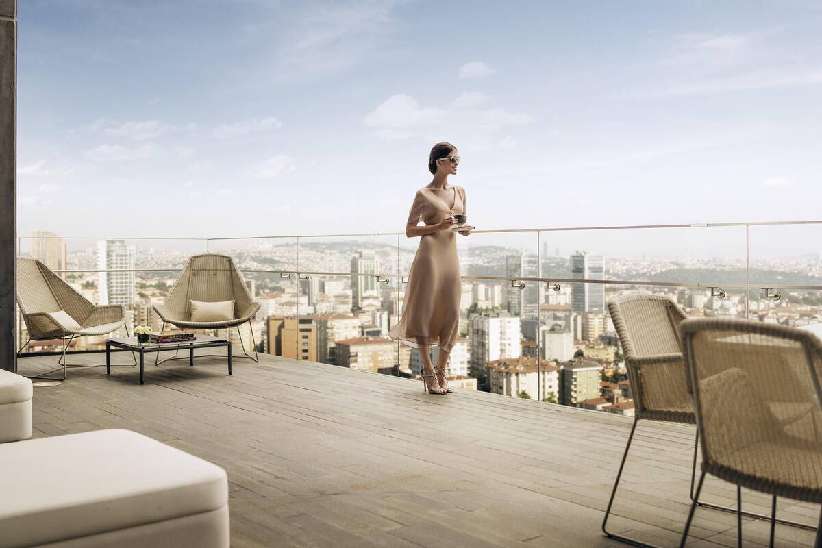 A woman standing on a balcony with a city view, two wicker chairs, and a table with books.
