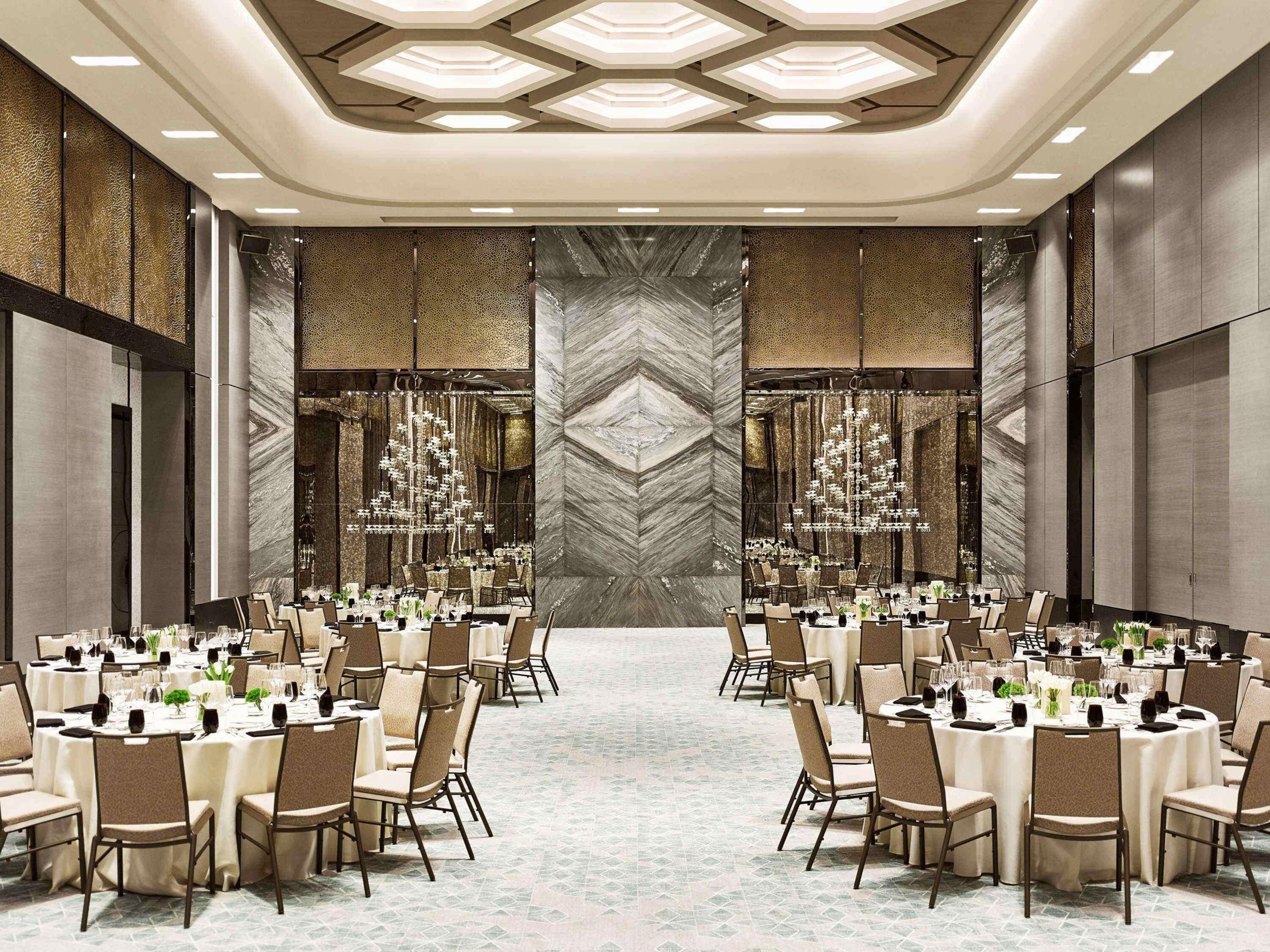 A large banquet hall with multiple round tables, high ceilings with geometric lighting, and a central marble wall.