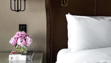 Una habitación de hotel con cabecera de madera, almohada blanca, mesita de noche con lámpara y un jarrón con flores rosas.