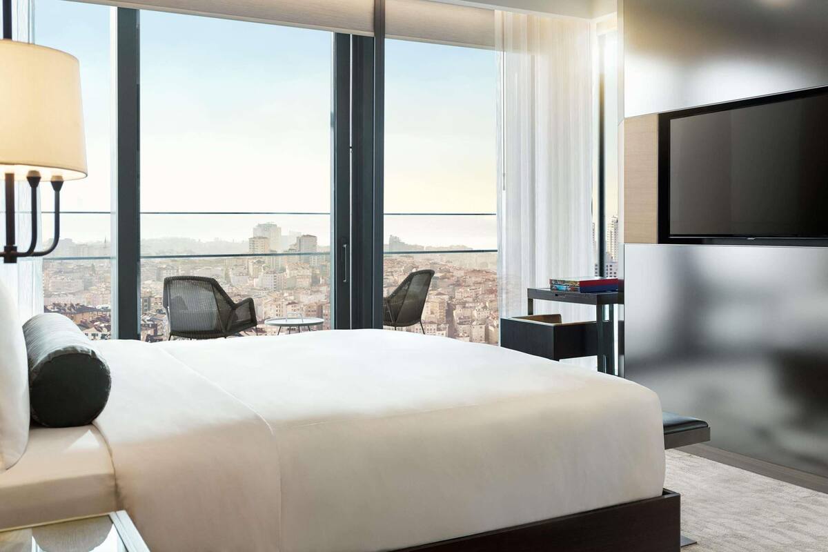 A modern hotel room with a large bed, a TV, and a city view through floor-to-ceiling windows.