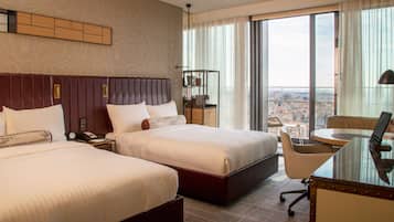 A hotel room with two beds, a desk, a chair, and a large window with a city view.