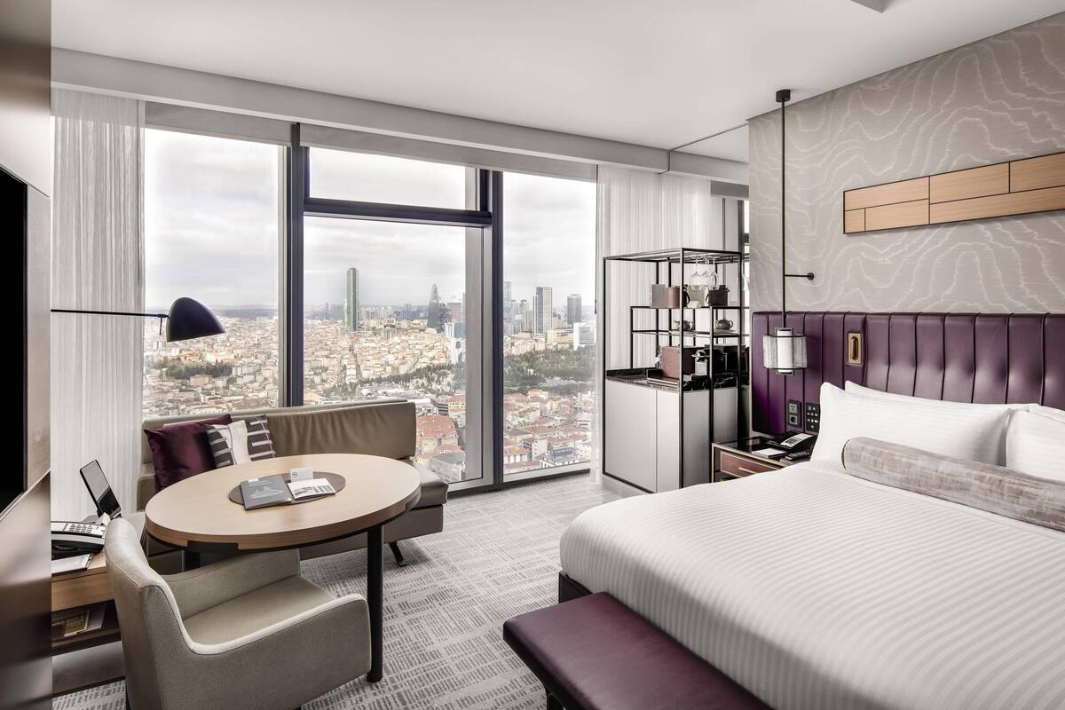 A modern hotel room with a large window offering a city view, a round dining table, a comfortable sofa, a neatly made bed, and a small side table with a lamp.