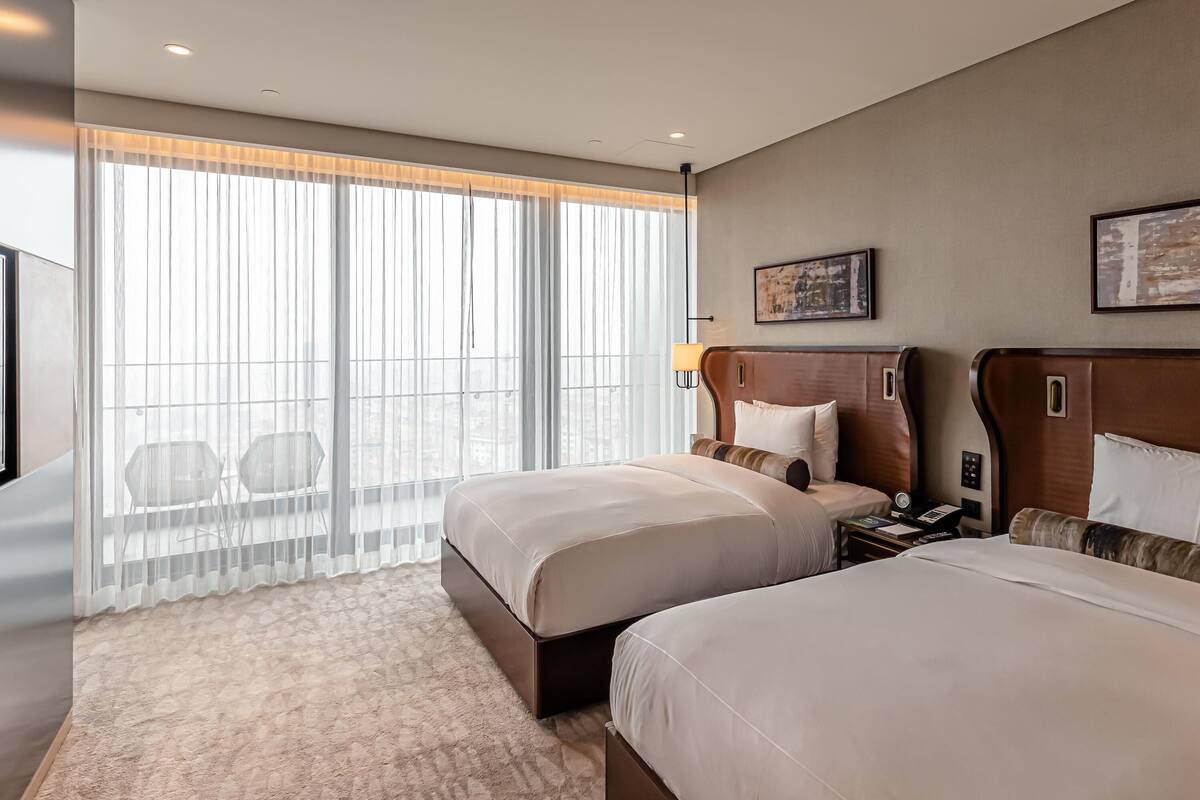 A hotel room with two beds, a large window with sheer curtains, a city view, and a painting on the wall.