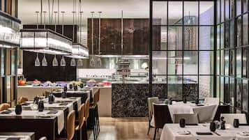 A modern restaurant interior with a central kitchen, dining tables, and chairs.