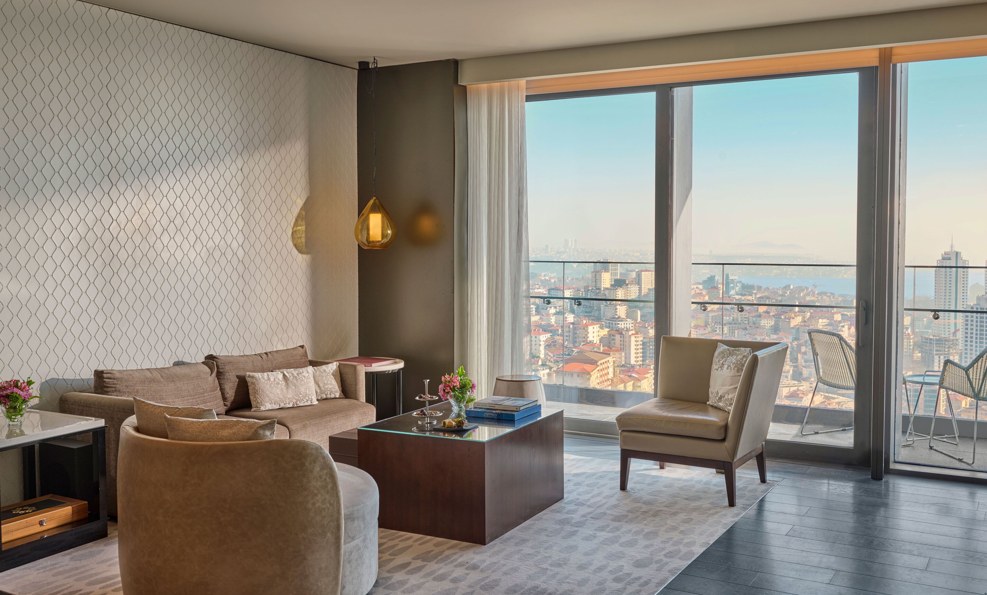 A modern living room with a sofa, armchairs, a coffee table, and a large window offering a city view.
