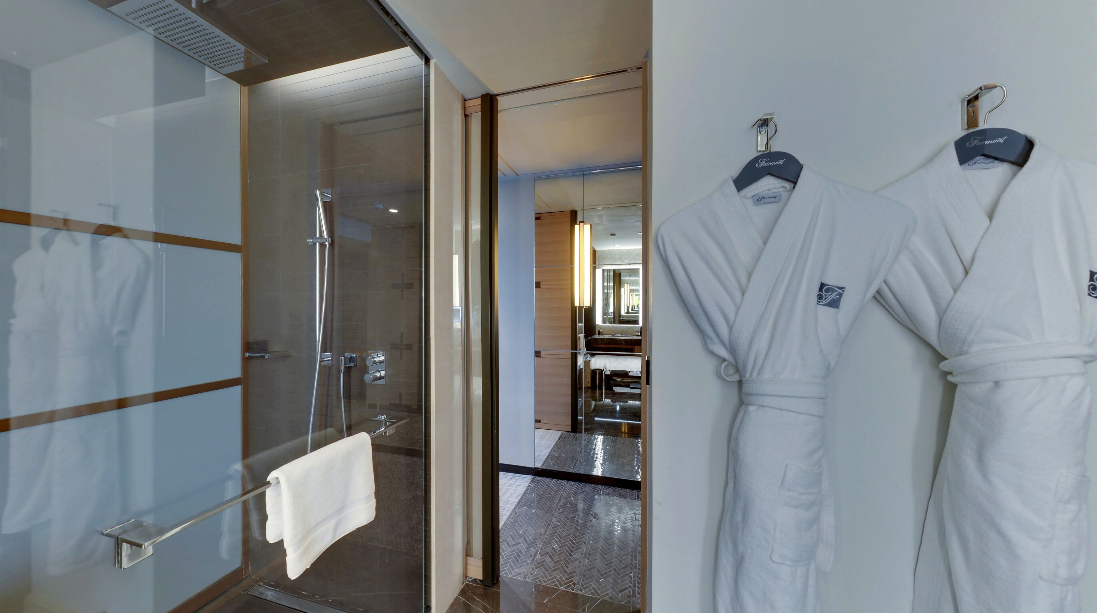 A hotel bathroom with a glass shower enclosure, a towel rack, and two white robes hanging on a rack.