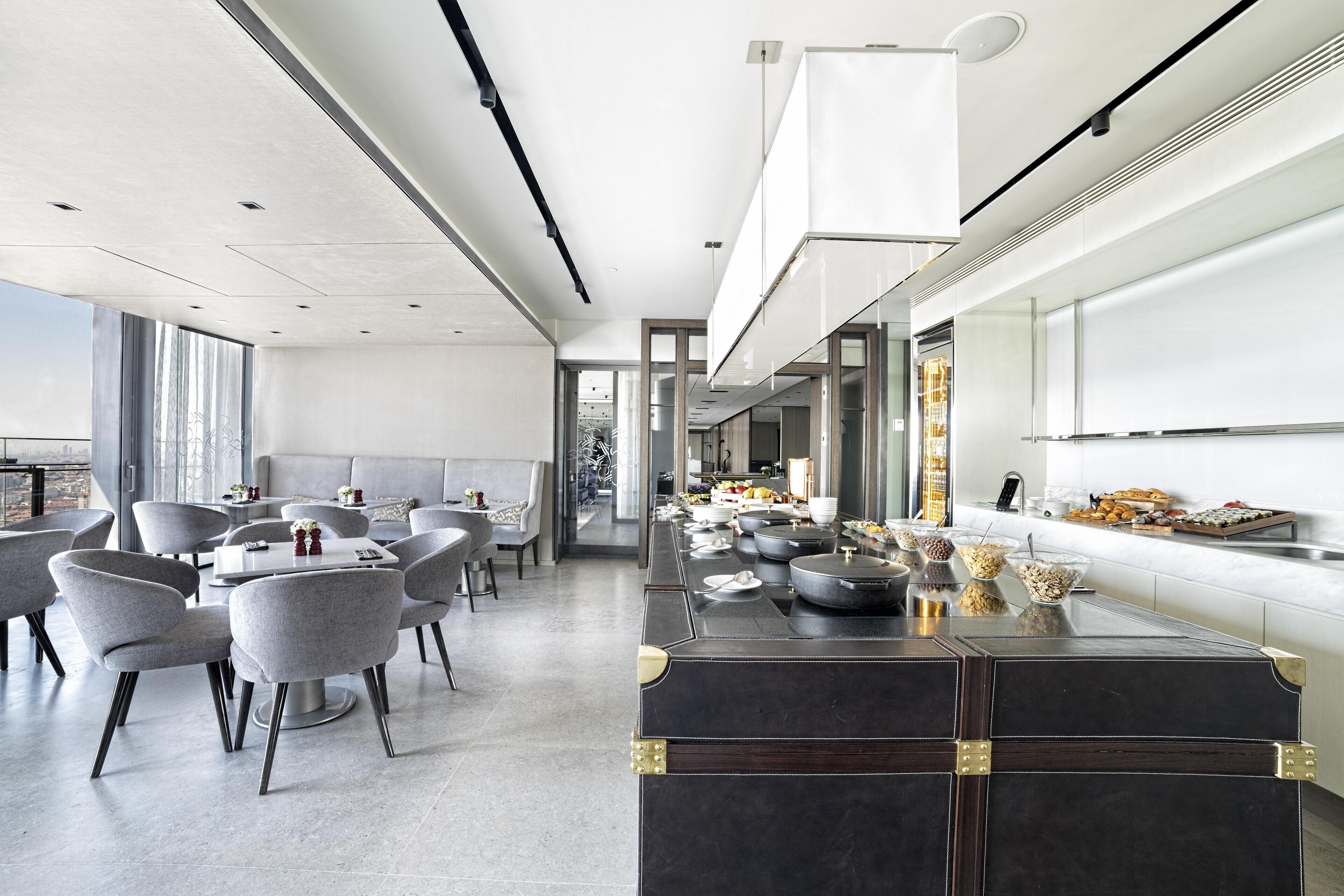 A modern dining area with a long counter, grey upholstered chairs, and a variety of food items displayed on the counter.