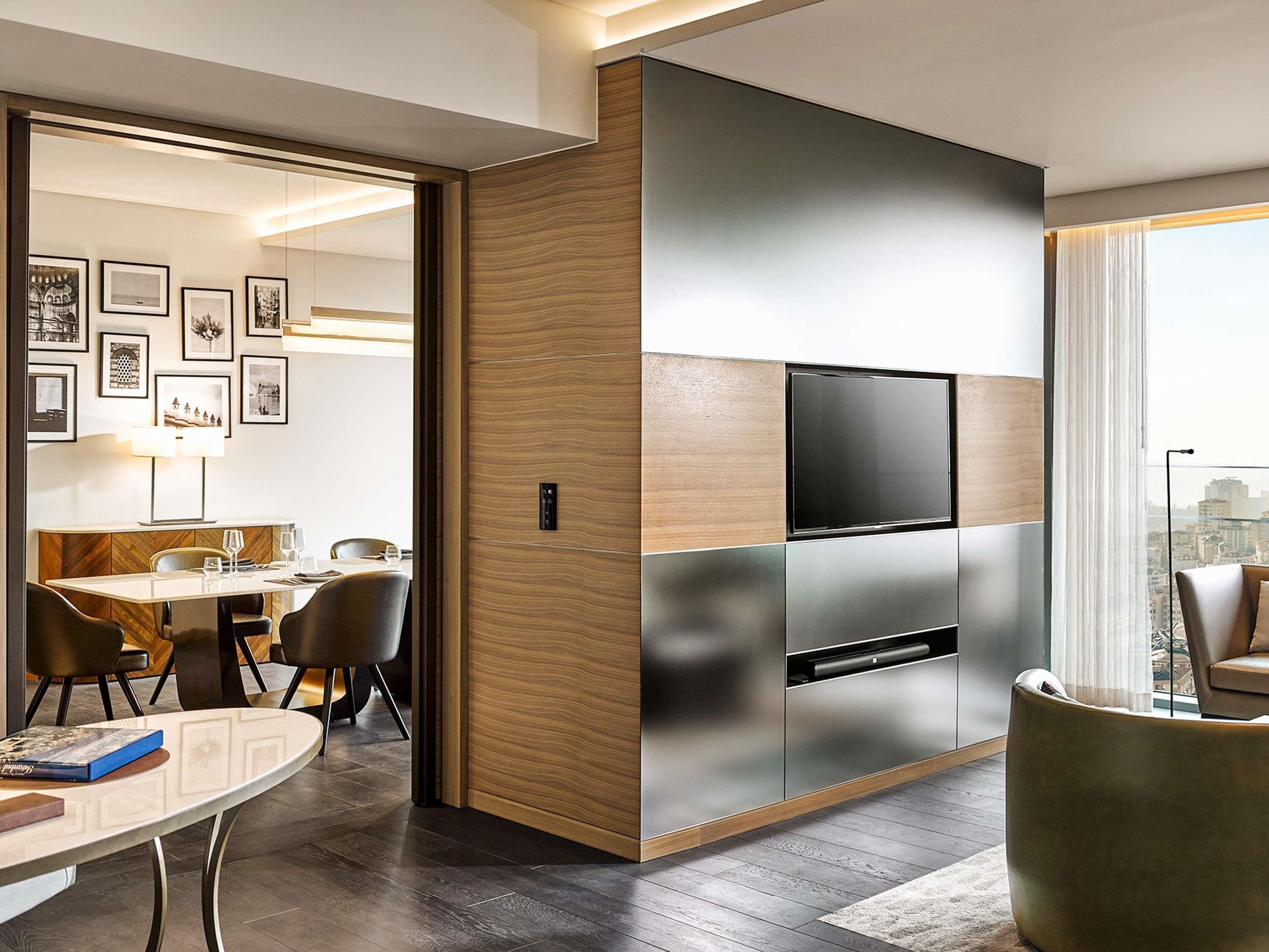 A modern hotel room with a flat-screen TV, a dining area, and a view of the city.