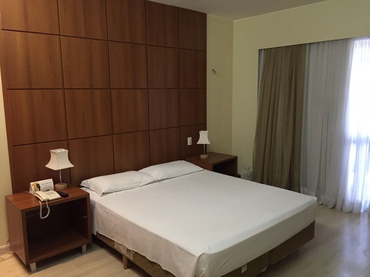 A hotel room with a large bed, wooden headboard, bedside tables with lamps, and a window with curtains.