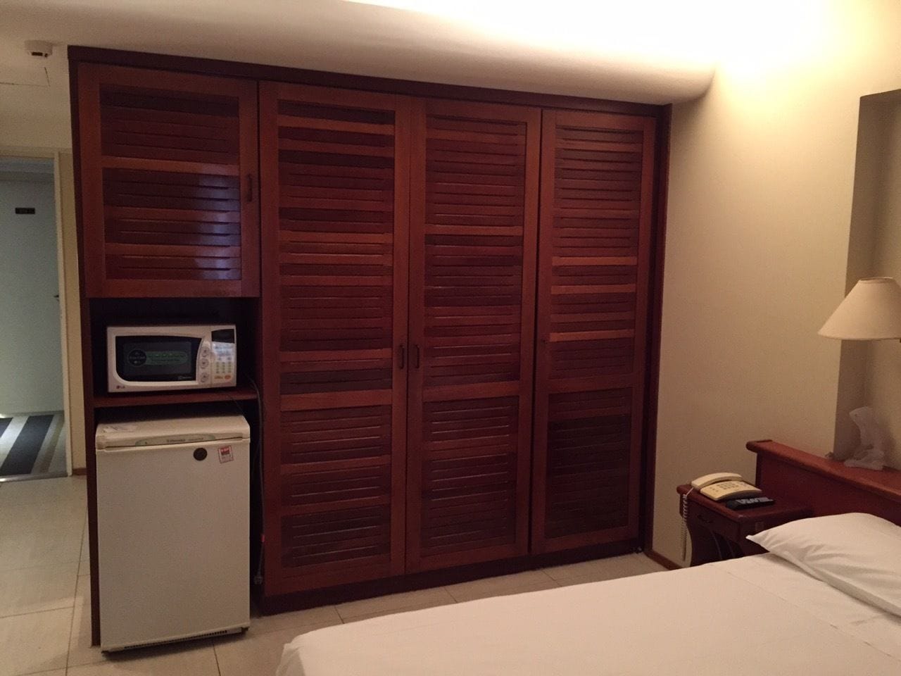 A hotel room with a large wooden wardrobe, a microwave, a mini-fridge, a bedside table with a lamp, and a bed.