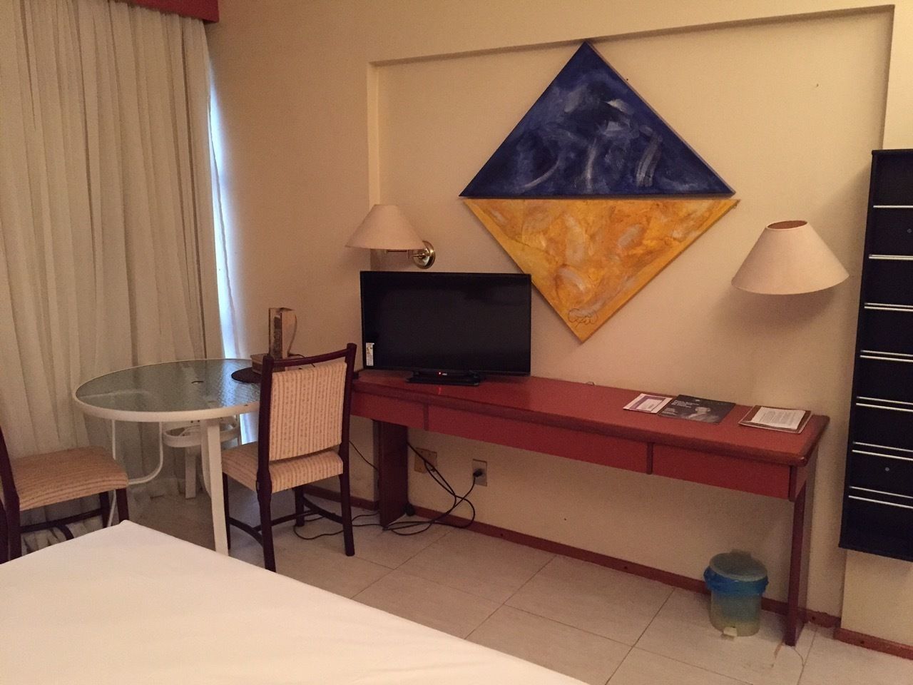 Una camera d'albergo con un letto, una scrivania con televisore, una sedia, un tavolino e un quadro appeso al muro.