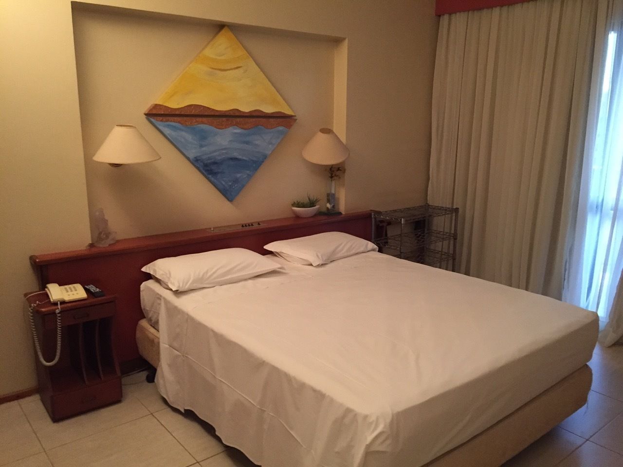 Une chambre d’hôtel avec un lit, deux lampes de chevet, un tableau au mur, un téléphone et une fenêtre avec des rideaux.