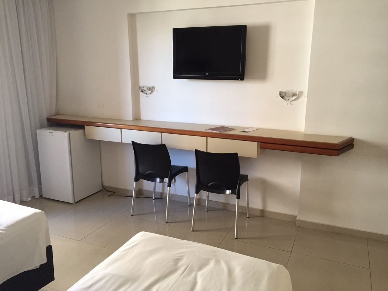 A hotel room with a bed, a desk with two chairs, a television mounted on the wall, and a small refrigerator.