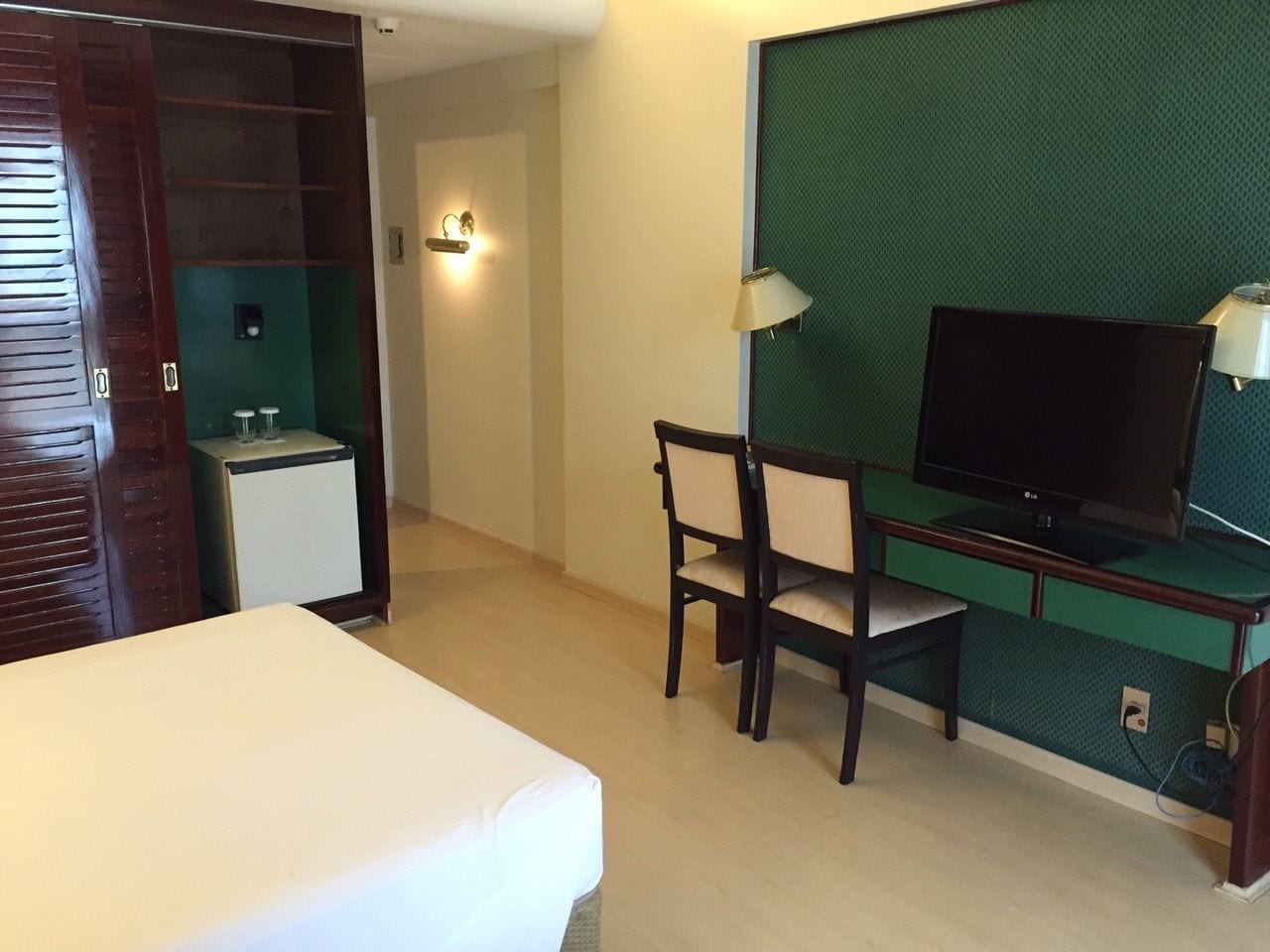 Quarto de hotel com cama, secretária com cadeira, televisão e pequeno frigorífico.