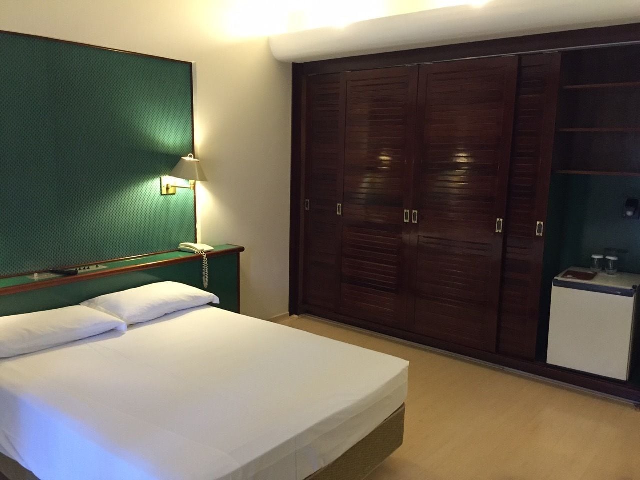 A hotel room with a bed, a nightstand, a lamp, a wardrobe, and a small refrigerator.