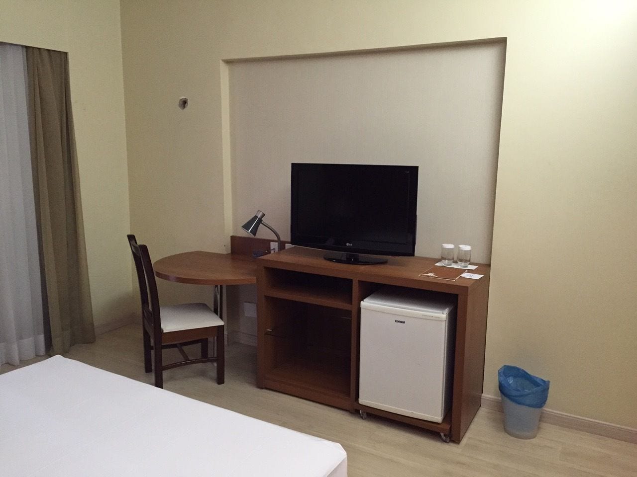 A hotel room with a bed, a wooden desk with a television, a small refrigerator, and a chair.