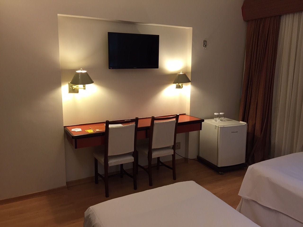 A hotel room with a desk, two chairs, a television, a minibar, and two beds.