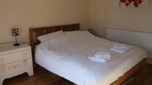 Un lit bien fait, avec du linge de lit et des serviettes blanches, une tête de lit en bois, une table de chevet avec une lampe et une table de nuit à motifs floraux.