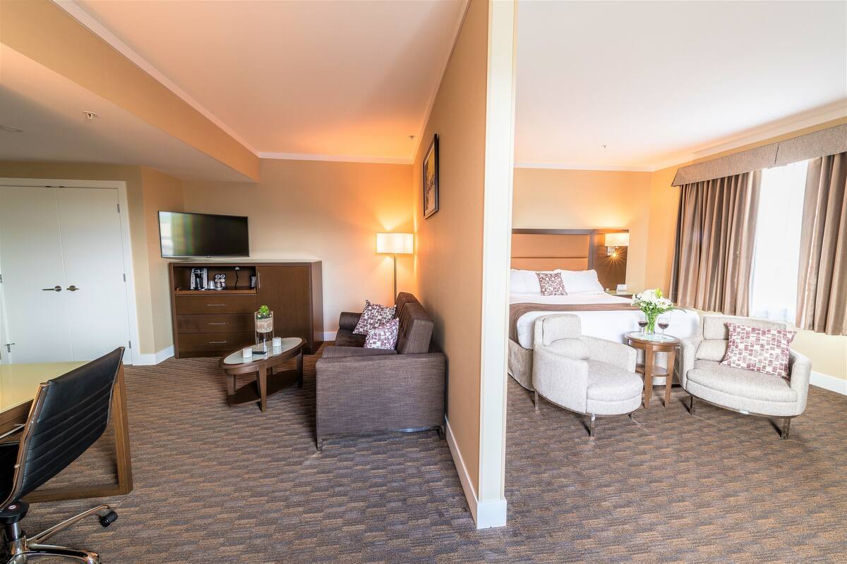 Habitación de hotel con sofá, dos sillones, escritorio, televisor, lámpara y cama.