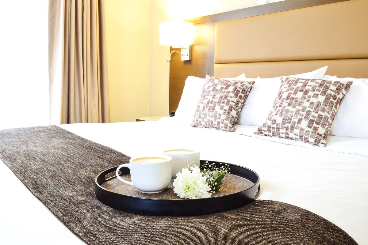 Una cama de hotel impecablemente arreglada con una bandeja que contiene dos tazas blancas y una pequeña flor blanca.