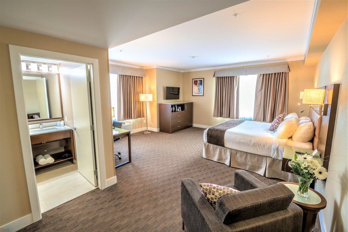 Habitación de hotel con cama, escritorio, silla, televisor y baño con espejo y toallero.