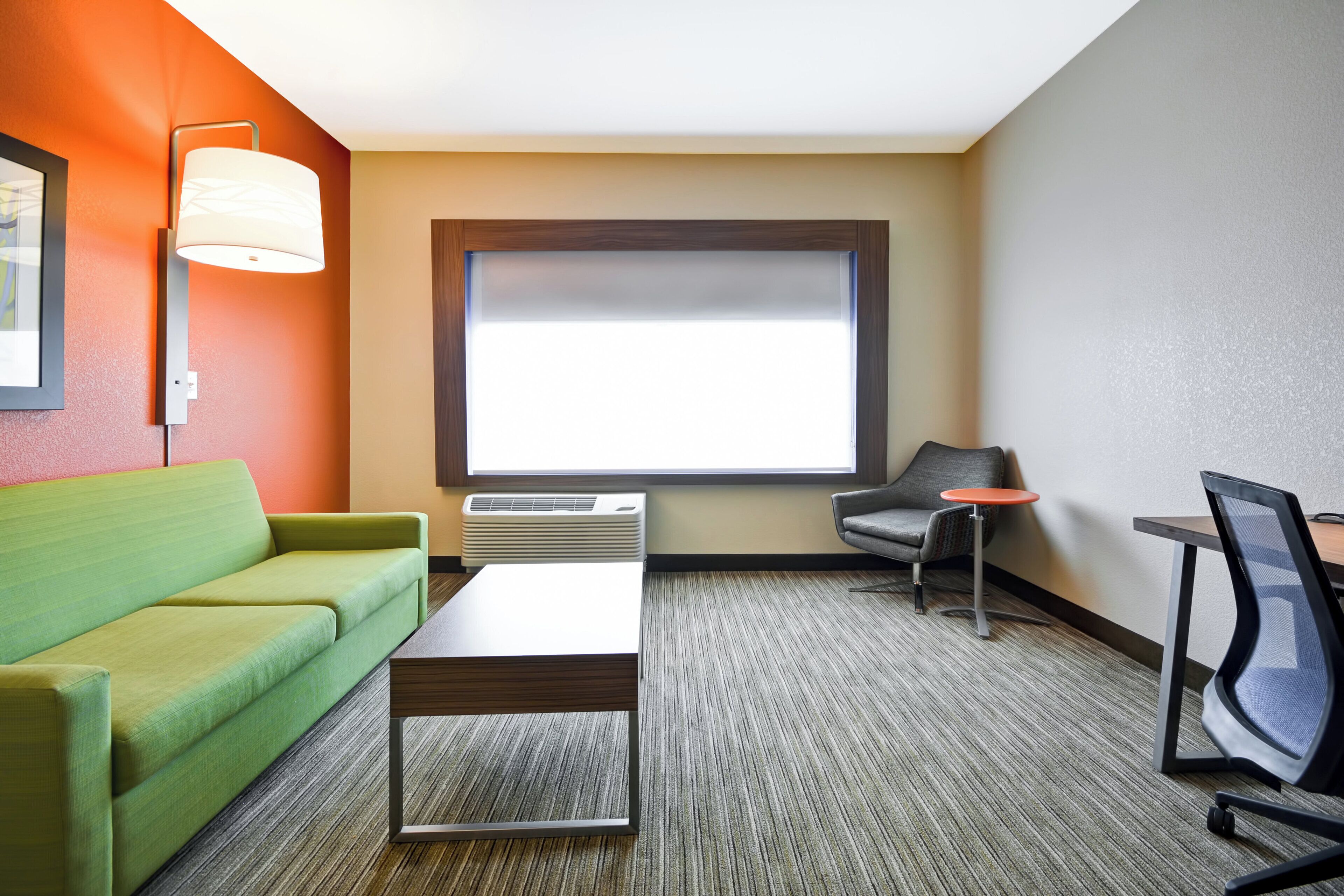 A hotel room with a green sofa, a small table, a grey armchair, a red side table, and a large window with blinds.