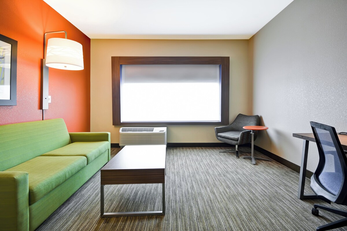 A hotel room with a green sofa, a small table, a grey armchair, a red side table, and a large window with blinds.