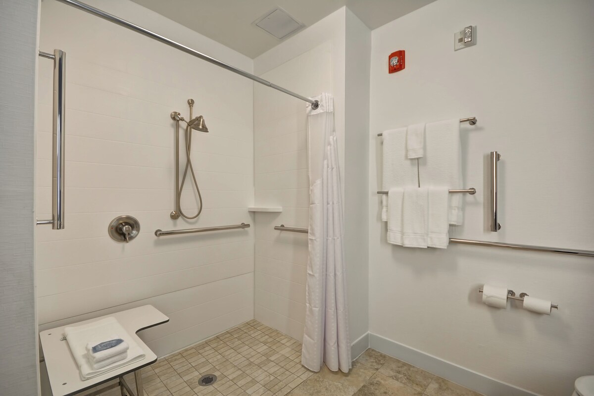 A modern bathroom with a walk-in shower, grab bars, a toilet with a paper towel holder, and a small sink with a towel.