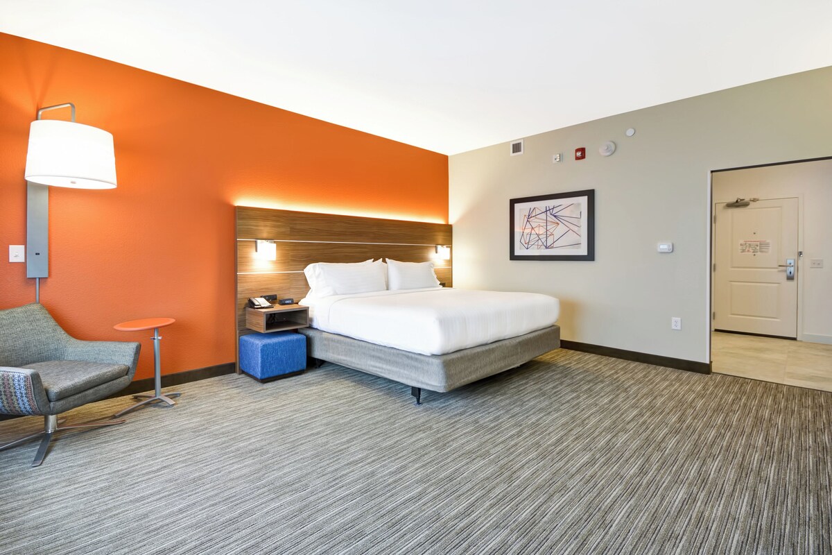 A hotel room with a bed, a desk, a chair, a lamp, and a door.